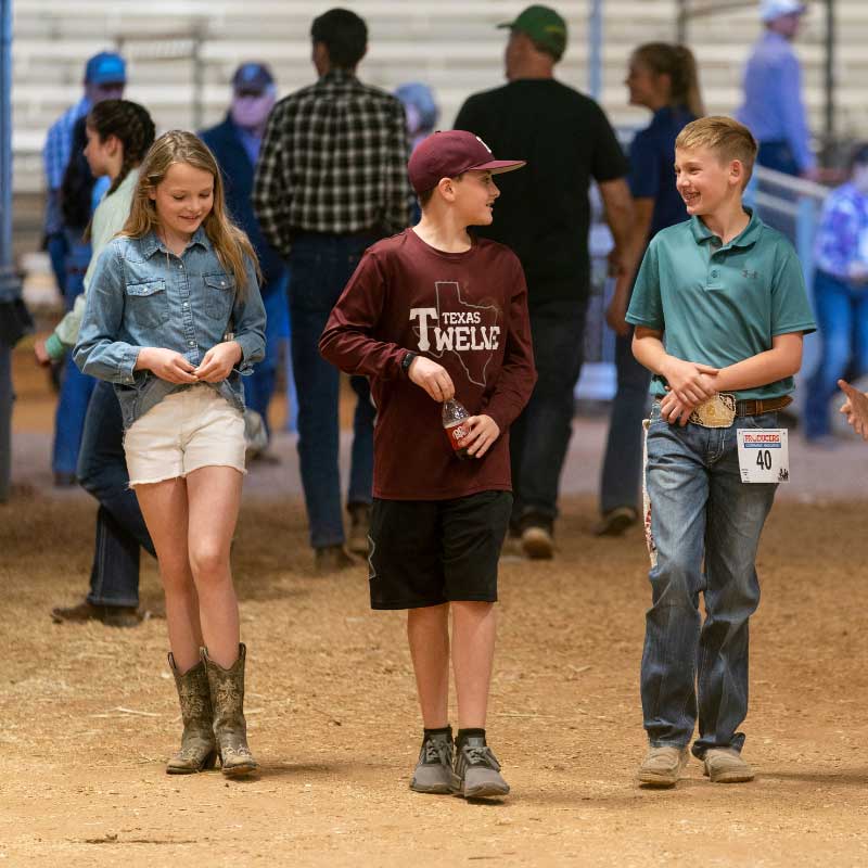 Three young people walk in an indoor area with a dirt floor, as they smile and talk.